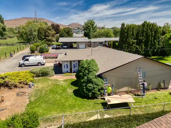 Aerial view of a tan ranch house with Top Line painters on ladders and a work table set up on the lawn