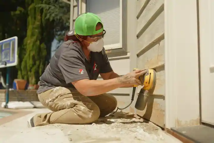 A Top Line painter sanding siding with an orbital sander, wearing a mask